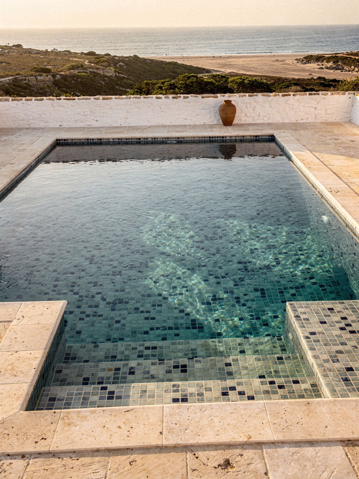 Pool mit blauem Glasmosaik und hellem Natursteinrand vor mediterraner Landschaft